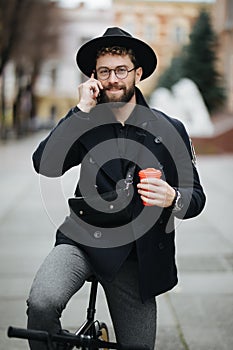 Smiling young stylish bearded hipster man using mobile phone while drinking coffee outdoors with bicycle