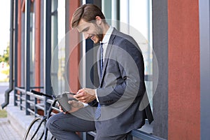 Smiling young stylish man using mobile phone while drinking coffee outdoors with bicycle.