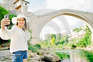Smiling young tourist making selfie with smartphone in front of historic landmark Old Bridge in Mostar, Bosnia and Herzegovina.