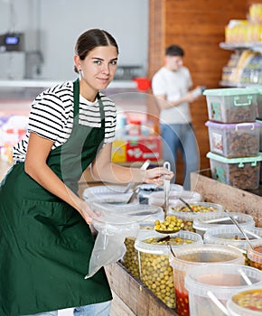 Smiling young vendoress pouring marinated olives into bag in grocery store