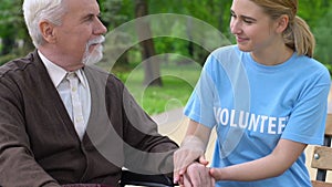 Smiling young volunteer supporting elderly disabled man in park, helping retiree