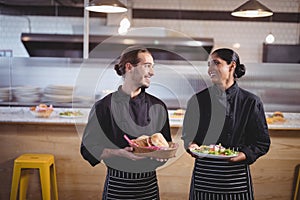 Smiling young wait staff looking at each other while holding fresh food in coffee shop