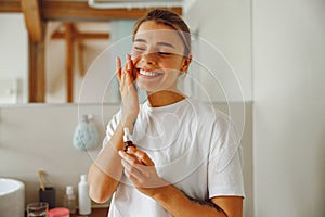Smiling young woman applying cosmetic serum onto her face in bathroom. Facial Skincare