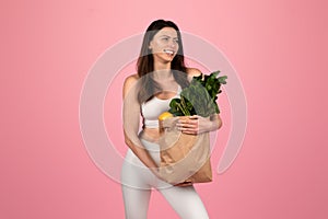 Smiling young woman in athletic wear holding a paper grocery bag full of fresh vegetables and fruits