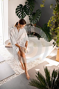 Smiling young woman in bathrobe dry brushing her legs with a natural wooden brush while sitting on the side of a bathtub