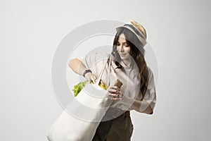 Smiling young woman in beige t-shirt and a hst with a mesh eco bag full of vegetables and on a white studio background