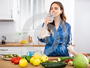 Smiling young woman in blue dressing gown drinking water in kitchen