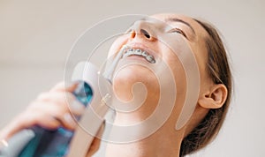 Smiling young woman with braces cleaning her teeth with oral irrigator, close-up.