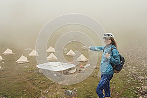 Smiling young woman in cap and blue sportswear with backpack hiker standing in green mountain valley and pointing with hand at