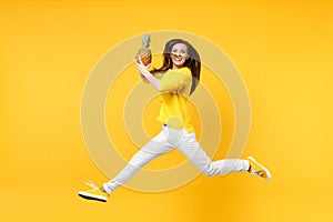 Smiling young woman in casual clothes jumping, hold fresh ripe pineapple fruit isolated on yellow orange wall background
