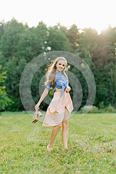 Smiling young woman in casual clothes strolls through the park. She is holding shoes in her hands. She is free and happy. The