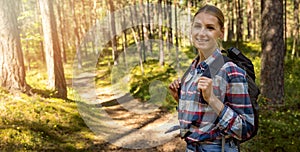 Smiling young woman in checkered shirt with backpack standing on forest trail. nature adventure hike. copy space