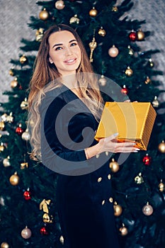 Smiling young woman with christmas present box near christmas tree in a black dress