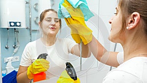 Smiling young woman cleaning and washing bathroom mirror with chemical detergent spray. Housewife doing home cleanup and