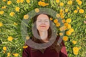 Smiling young woman with closed eyes relaxing on a meadow with many dandelions in the spring sun. Top down view.