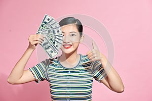 Smiling young woman closes half of her face with a fan of banknotes cash and shows thumb up on pink background