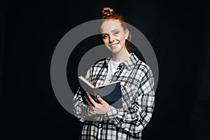 Smiling young woman college student holding opened book and looking at camera on black background