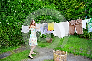 Smiling young woman in colorful dress hanging laundry on clothesline at the backyard