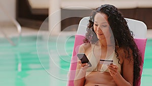 Smiling young woman customer holding credit card and smartphone sitting in lawn chair. Happy female shopper in swimsuit