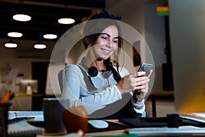 Smiling young woman designer sitting in office working using mobile phone at night.