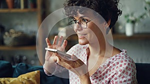 Smiling young woman is dialing telephone number then talking in loudspeaker mode holding smartphone in hand. Modern