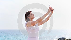 Smiling young woman doing selfie photo on cellphone against blue Mediterranean sea in slow motion