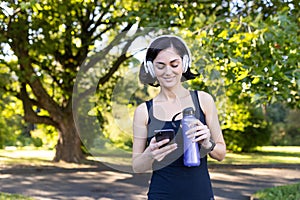 Smiling young woman doing sports and jogging outdoors in park, standing resting in headphones, holding water bottle and