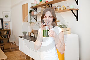 Smiling young woman drinking green smoothie juice in kitchen.