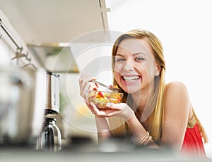 Smiling young woman eating fresh fruits salad in kitchen