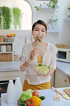 Smiling young woman eating fresh salad in modern kitchen