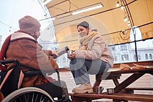 Smiling young woman enjoying time with man in wheelchair sharing hot drinks in winter city