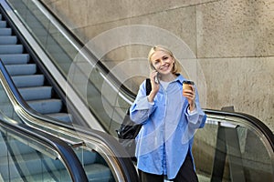 Smiling young woman on escalator, talking on mobile phone, drinking coffee and looking happy, going to university or