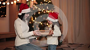 Smiling young woman exchanging sharing xmas gifts with her cute preschool daughter in santa hats