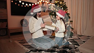 Smiling young woman exchanging sharing xmas gifts with her cute preschool daughter in santa hats