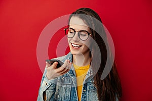 Smiling young woman in eyeglasses holding phone speak activate virtual digital voice recognition assistant on smartphone