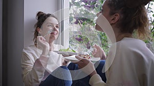 Smiling young woman feeding twin sister with healthful salad sitting on comfortable windowsill at home. Portrait of