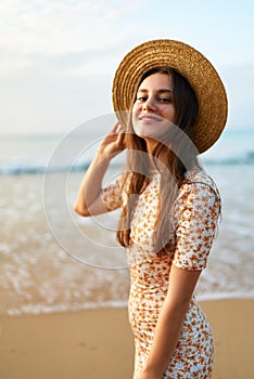 Smiling young woman in floral dress and straw hat stands on sandy beach. Fashionable female enjoys summer by sea at