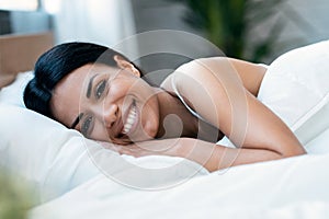 Smiling young woman with fresh skin in underwear having fun lying on bed while looking at camera at home