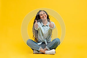 Smiling young woman gesturing thumbs up with both hands, recommending something on yellow studio background
