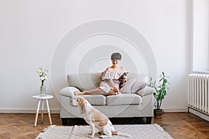 Smiling young woman in glasses looking with smile at beagle dog sitting on carpet beside sofa. Indoor portrait of lovely