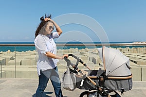 Smiling young woman, a happy mother, pushing a baby stroller while walking along the embankment of a resort town