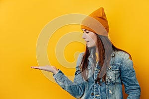 Smiling young woman in hat looks pleasant to her palm hand, showing something in her hand, dressed in denim jacket