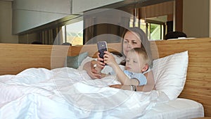 Smiling young woman with her child taking a selfie on the phone lying on the bed