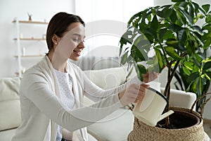Smiling young woman holding can, watering green houseplant at home
