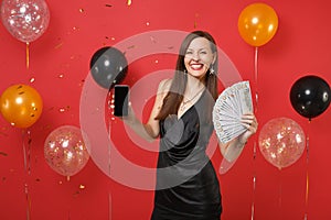 Smiling young woman holding mobile phone with blank empty black screen, bundle lots of dollars, cash money on bright red