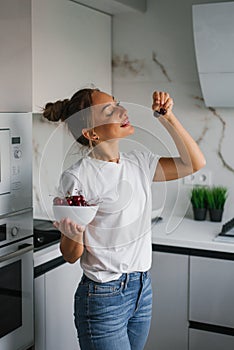Smiling young woman holds a plate of fresh cherry berries in her hand in the kitchen and wants to eat it