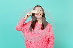 Smiling young woman in knitted pink sweater looking up, covering eye with credit card in hand isolated on blue turquoise