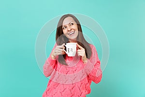 Smiling young woman in knitted pink sweater looking up and hold in hands cup of coffee or tea isolated on blue turquoise