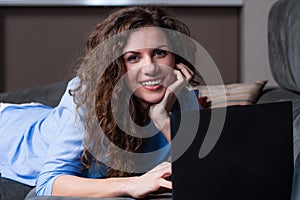 Smiling young woman laying on couch and using laptop.