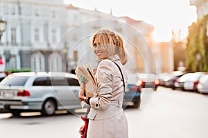 Smiling young woman in light-brown trench coat spending time outdoor after work. Photo of cheerful girl with blone hair